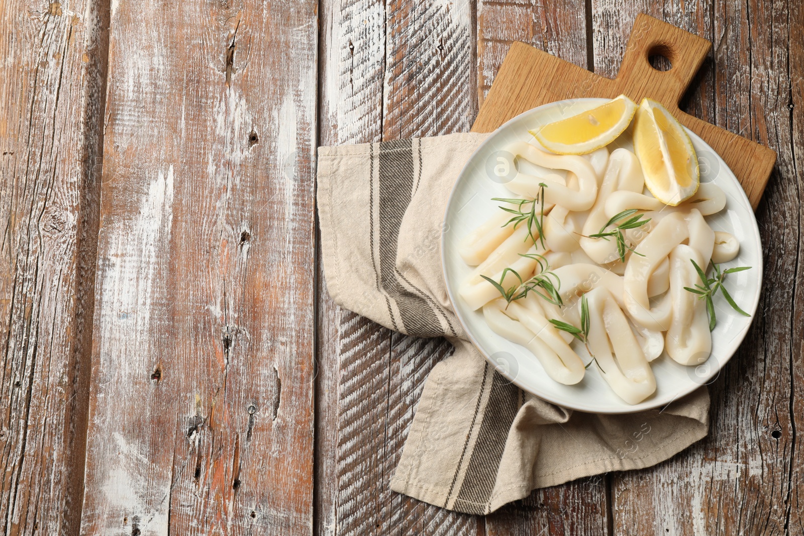 Photo of Uncooked squid rings with lemon slices and rosemary on wooden table, top view. Space for text