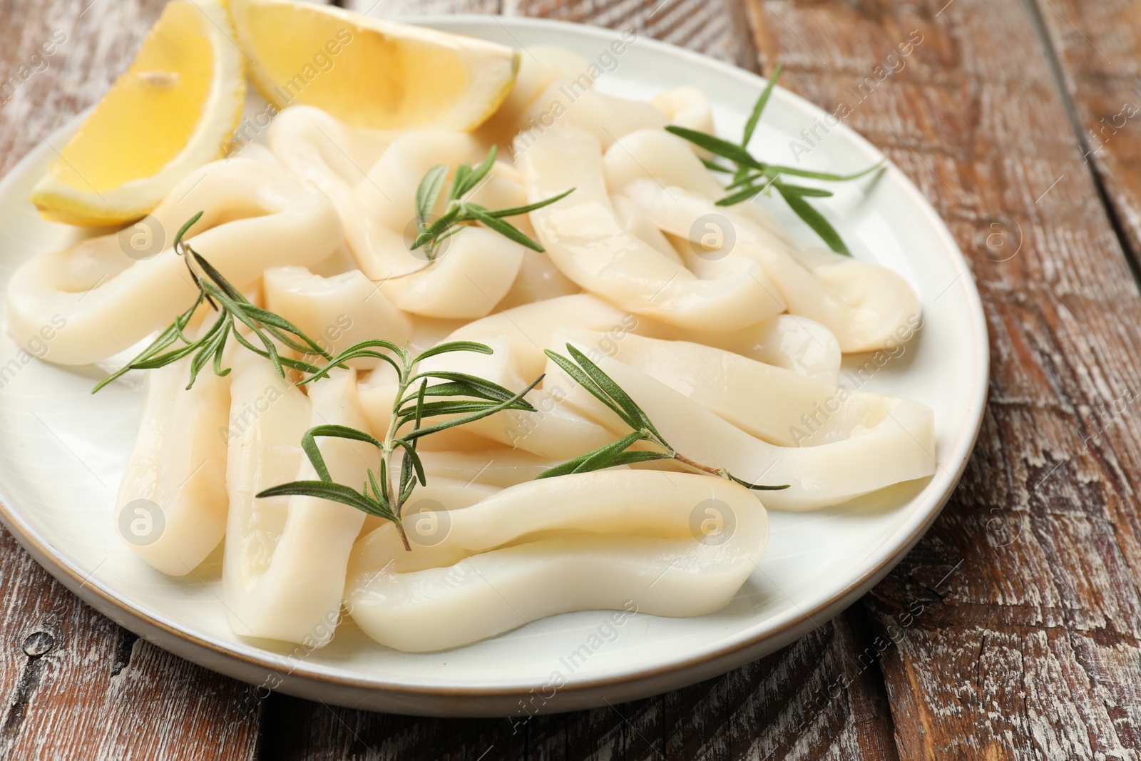 Photo of Uncooked squid rings with lemon slices and rosemary on wooden table, closeup
