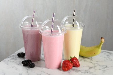 Photo of Milkshakes of different flavors in plastic cups and ingredients on white marble table, closeup