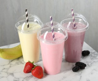 Photo of Milkshakes of different flavors in plastic cups and ingredients on white marble table, closeup