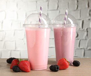 Photo of Tasty milkshakes in plastic cups and fresh berries on wooden table, closeup