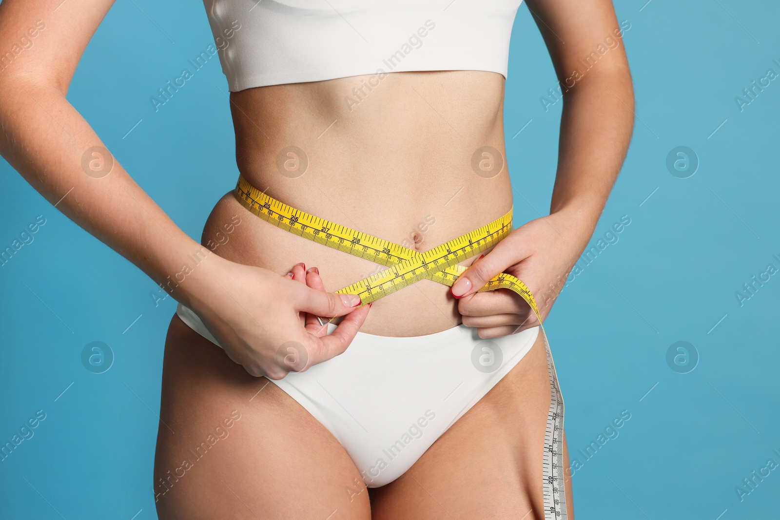 Photo of Young woman measuring her flat belly with tape on light blue background, closeup