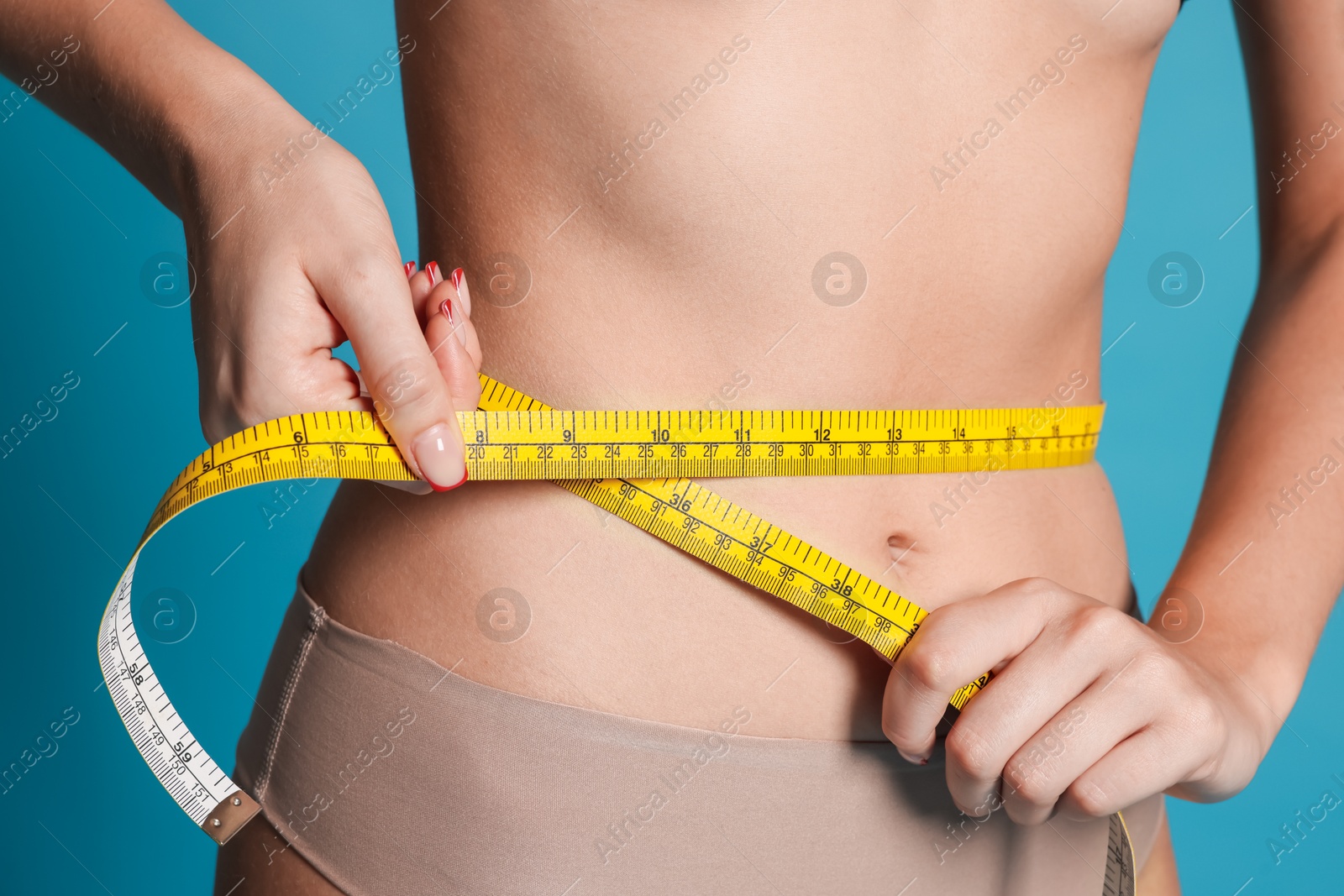 Photo of Young woman measuring her flat belly with tape on light blue background, closeup