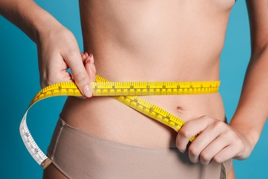 Photo of Young woman measuring her flat belly with tape on light blue background, closeup