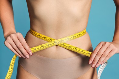 Photo of Young woman measuring her flat belly with tape on light blue background, closeup