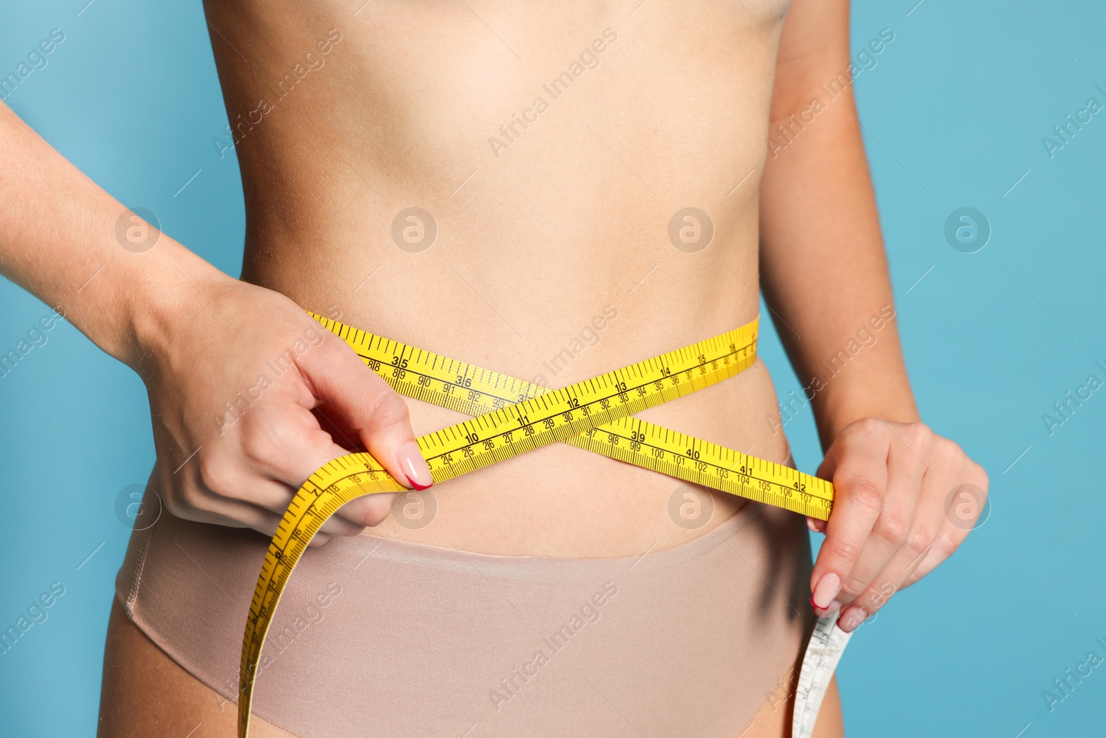 Photo of Young woman measuring her flat belly with tape on light blue background, closeup