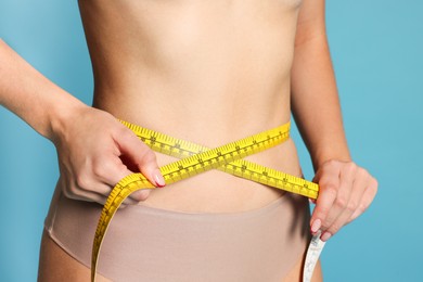 Photo of Young woman measuring her flat belly with tape on light blue background, closeup