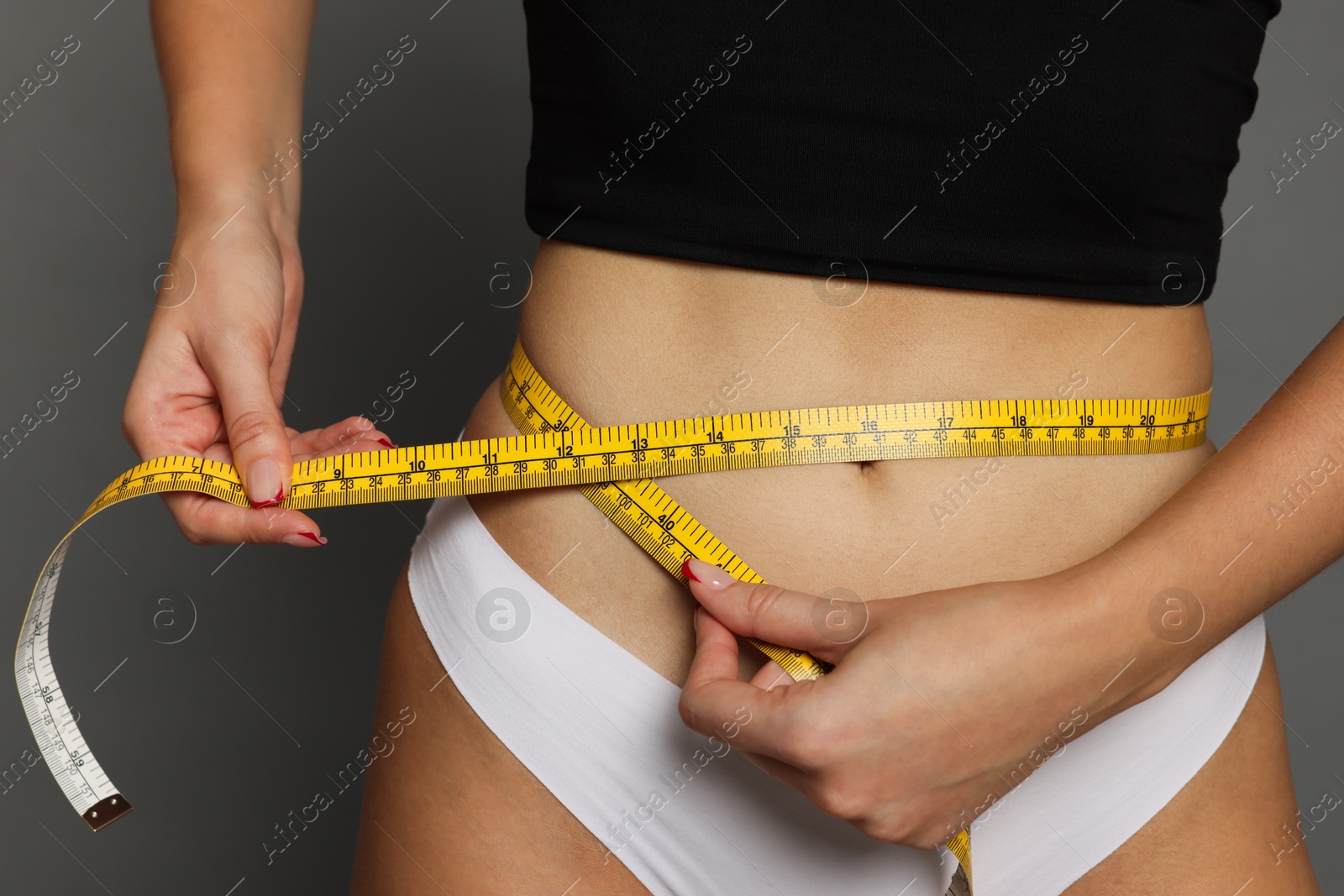 Photo of Young woman measuring her flat belly with tape on grey background, closeup