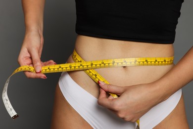 Photo of Young woman measuring her flat belly with tape on grey background, closeup
