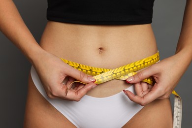 Photo of Young woman measuring her flat belly with tape on grey background, closeup