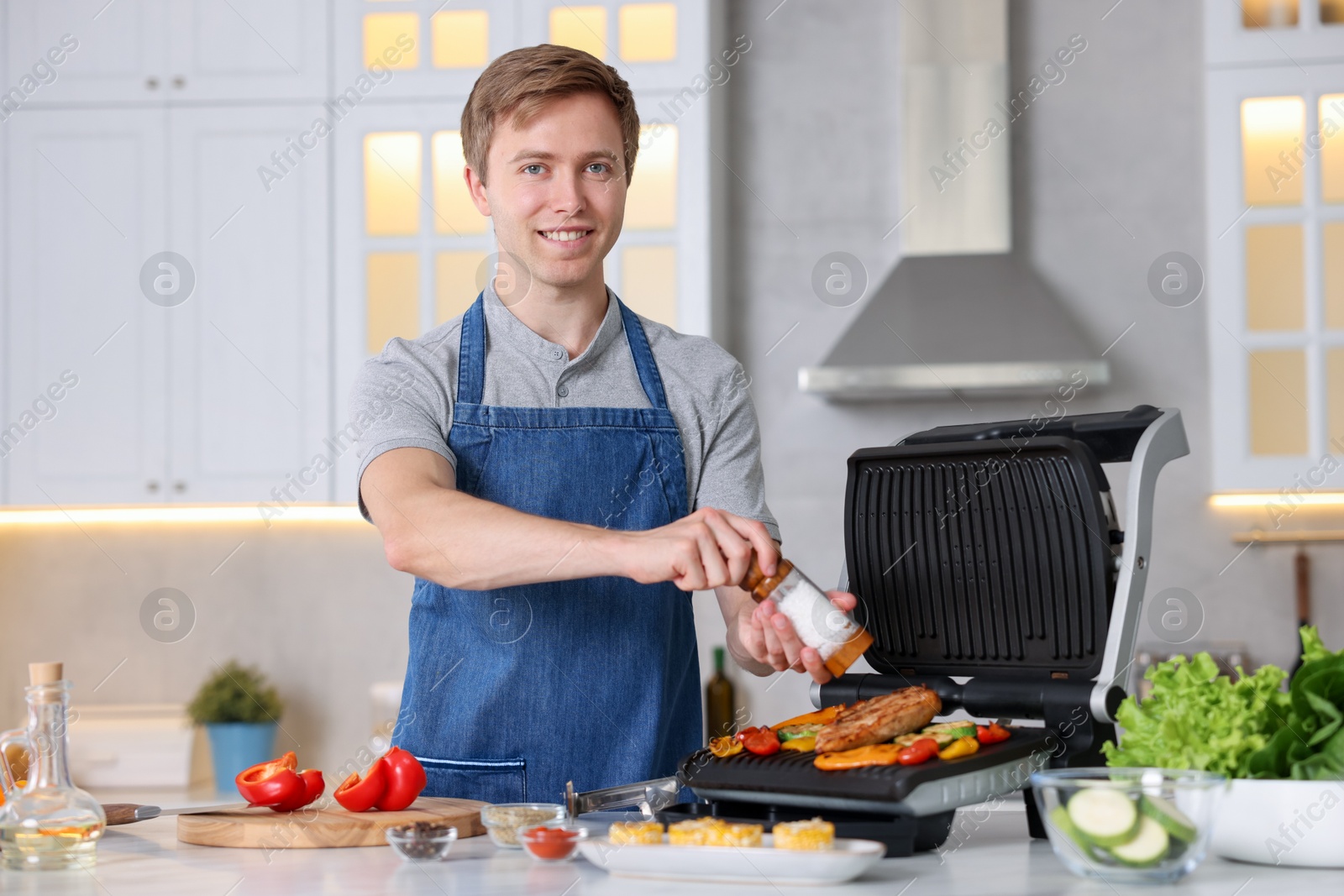 Photo of Man adding salt to vegetables and meat cooking on electric grill in kitchen