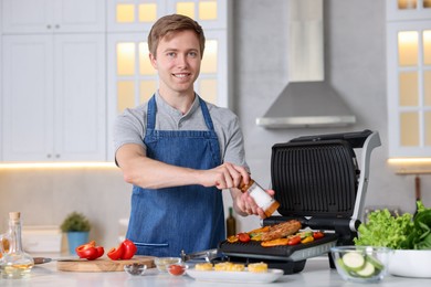 Photo of Man adding salt to vegetables and meat cooking on electric grill in kitchen
