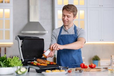Photo of Man adding salt to vegetables and meat cooking on electric grill in kitchen
