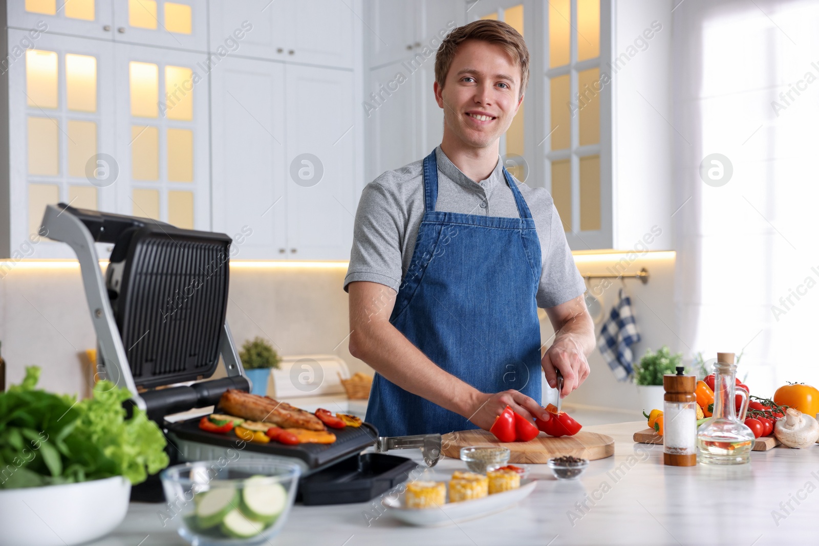 Photo of Man cutting pepper while cooking meat and vegetables on electric grill in kitchen