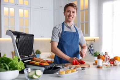 Photo of Man cutting pepper while cooking meat and vegetables on electric grill in kitchen