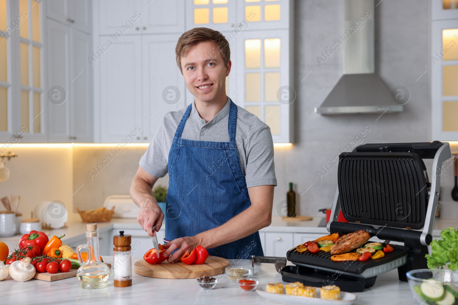 Photo of Man cutting pepper while cooking meat and vegetables on electric grill in kitchen