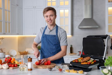 Photo of Man cutting pepper while cooking meat and vegetables on electric grill in kitchen