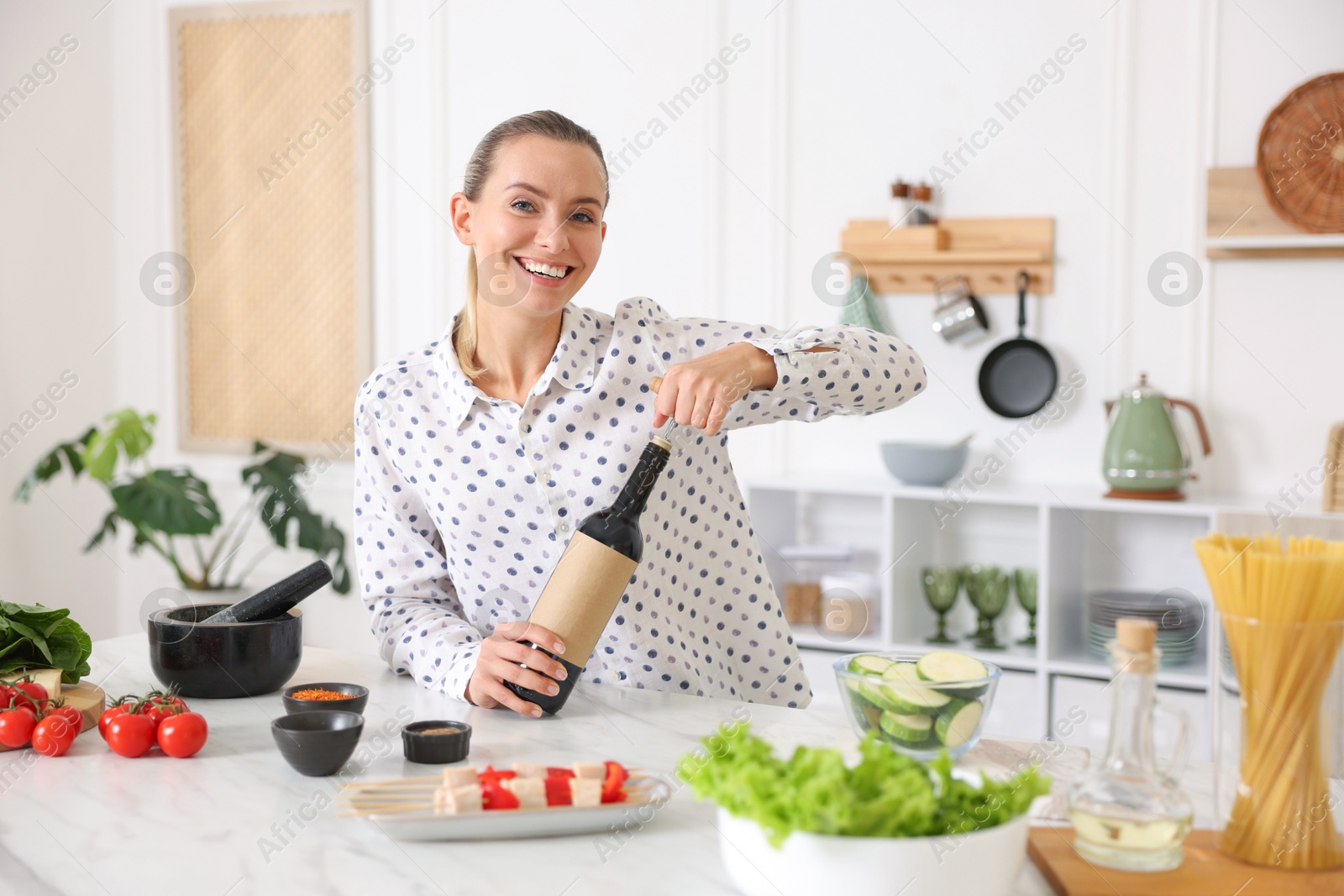 Photo of Smiling woman opening bottle of wine while cooking at table in kitchen