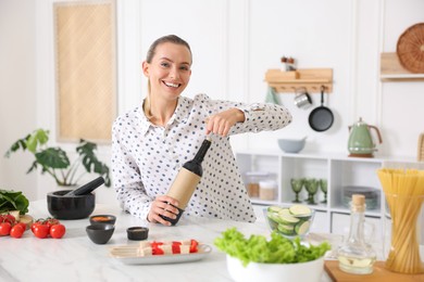 Photo of Smiling woman opening bottle of wine while cooking at table in kitchen