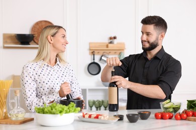 Photo of Smiling man opening bottle of wine while cooking with his wife at table in kitchen