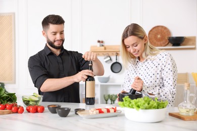 Photo of Smiling man opening bottle of wine while cooking with his wife at table in kitchen