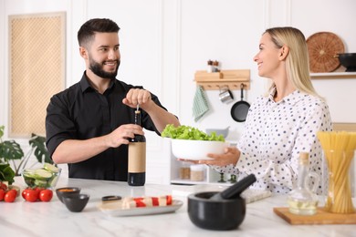 Photo of Smiling man opening bottle of wine while cooking with his wife at table in kitchen