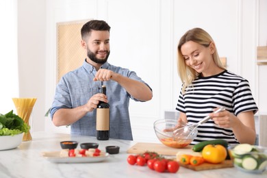 Photo of Smiling man opening bottle of wine while cooking with his wife at table in kitchen