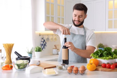 Photo of Smiling man opening bottle of wine while cooking at table in kitchen
