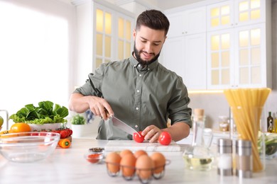 Photo of Cooking process. Smiling man cutting tomato at table in kitchen
