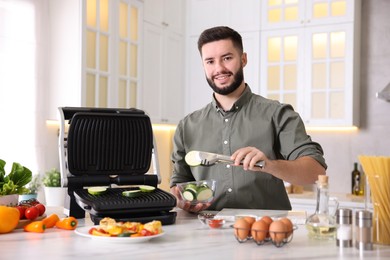 Photo of Smiling man cooking zucchini on electric grill at table in kitchen