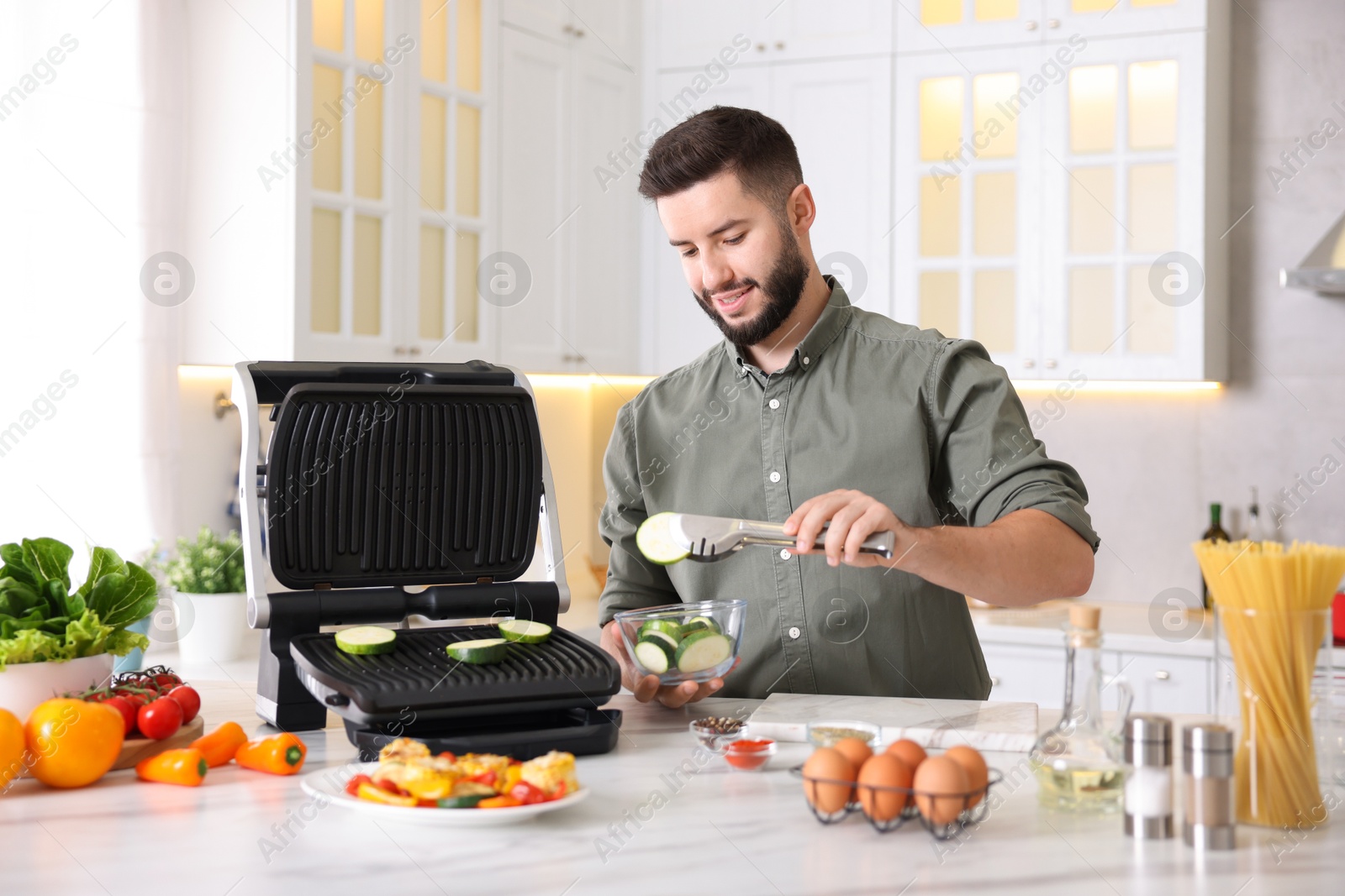 Photo of Smiling man cooking zucchini on electric grill at table in kitchen