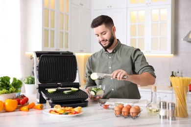 Photo of Smiling man cooking zucchini on electric grill at table in kitchen