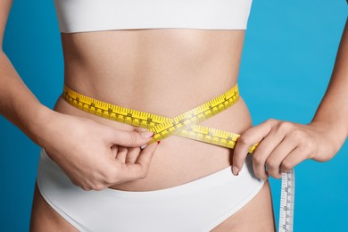 Photo of Young woman measuring her flat belly with tape on light blue background, closeup