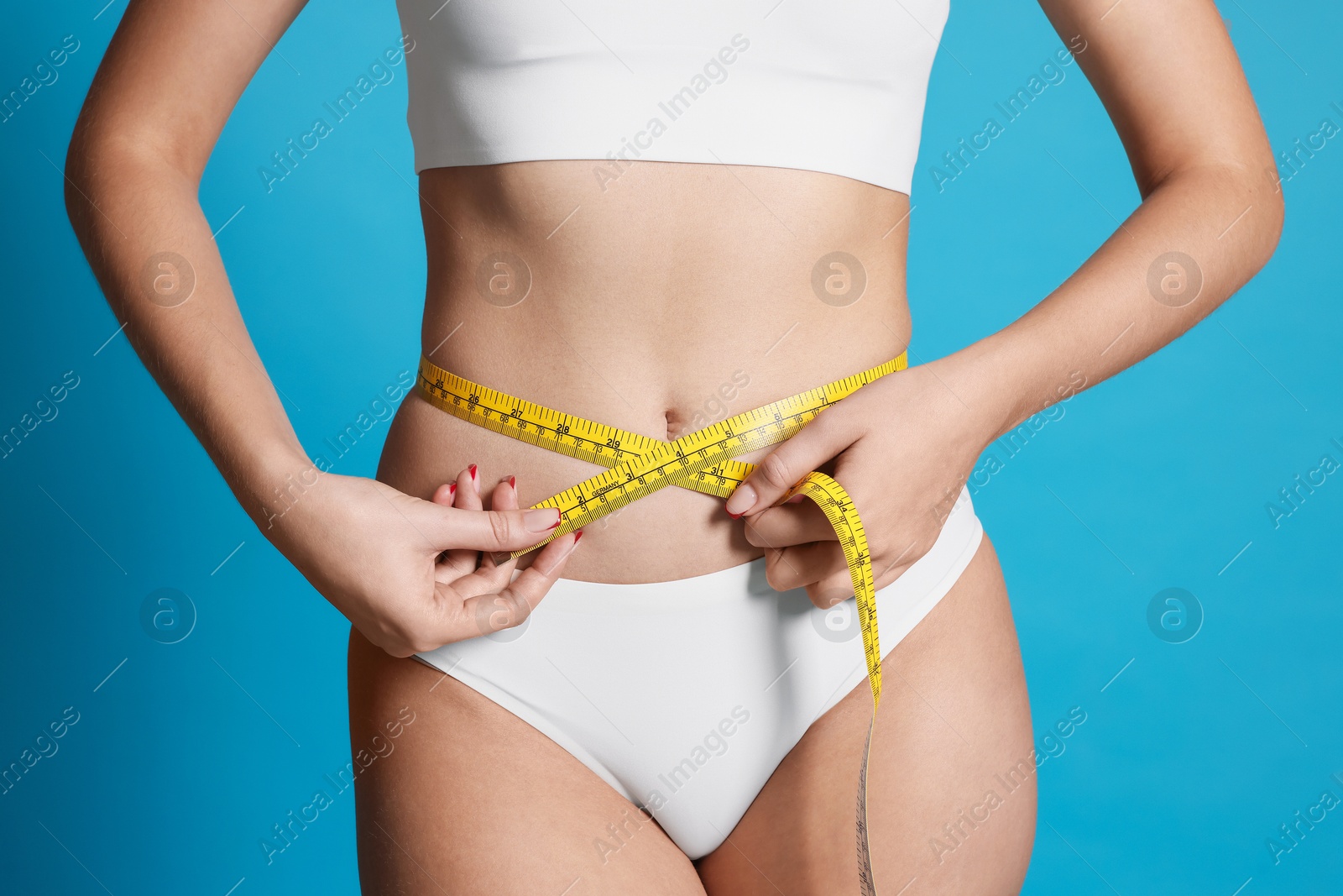 Photo of Young woman measuring her flat belly with tape on light blue background, closeup