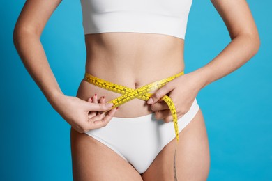 Photo of Young woman measuring her flat belly with tape on light blue background, closeup