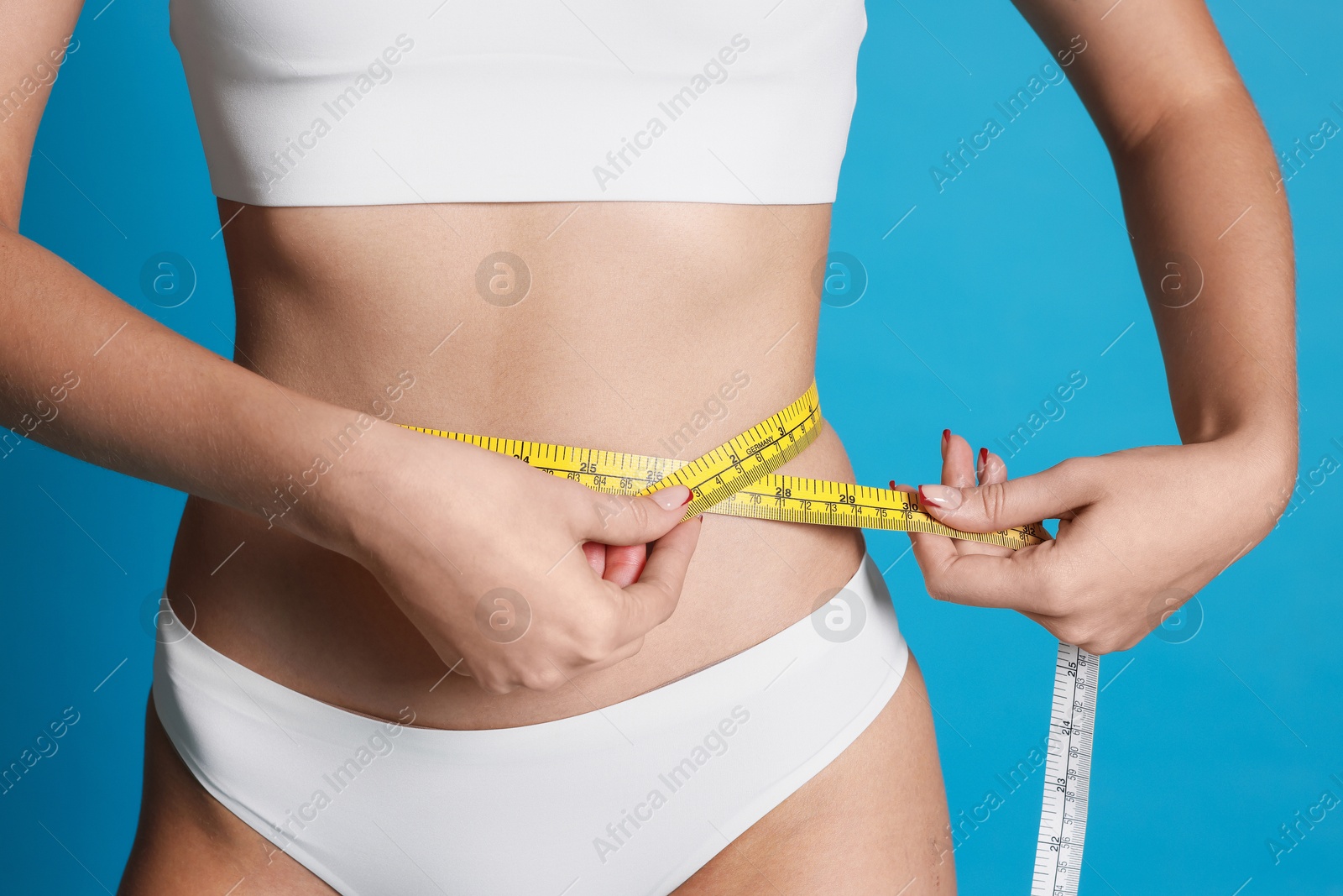 Photo of Young woman measuring her flat belly with tape on light blue background, closeup