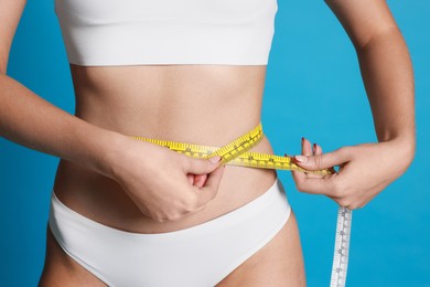 Photo of Young woman measuring her flat belly with tape on light blue background, closeup