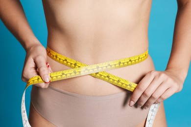 Photo of Young woman measuring her flat belly with tape on light blue background, closeup
