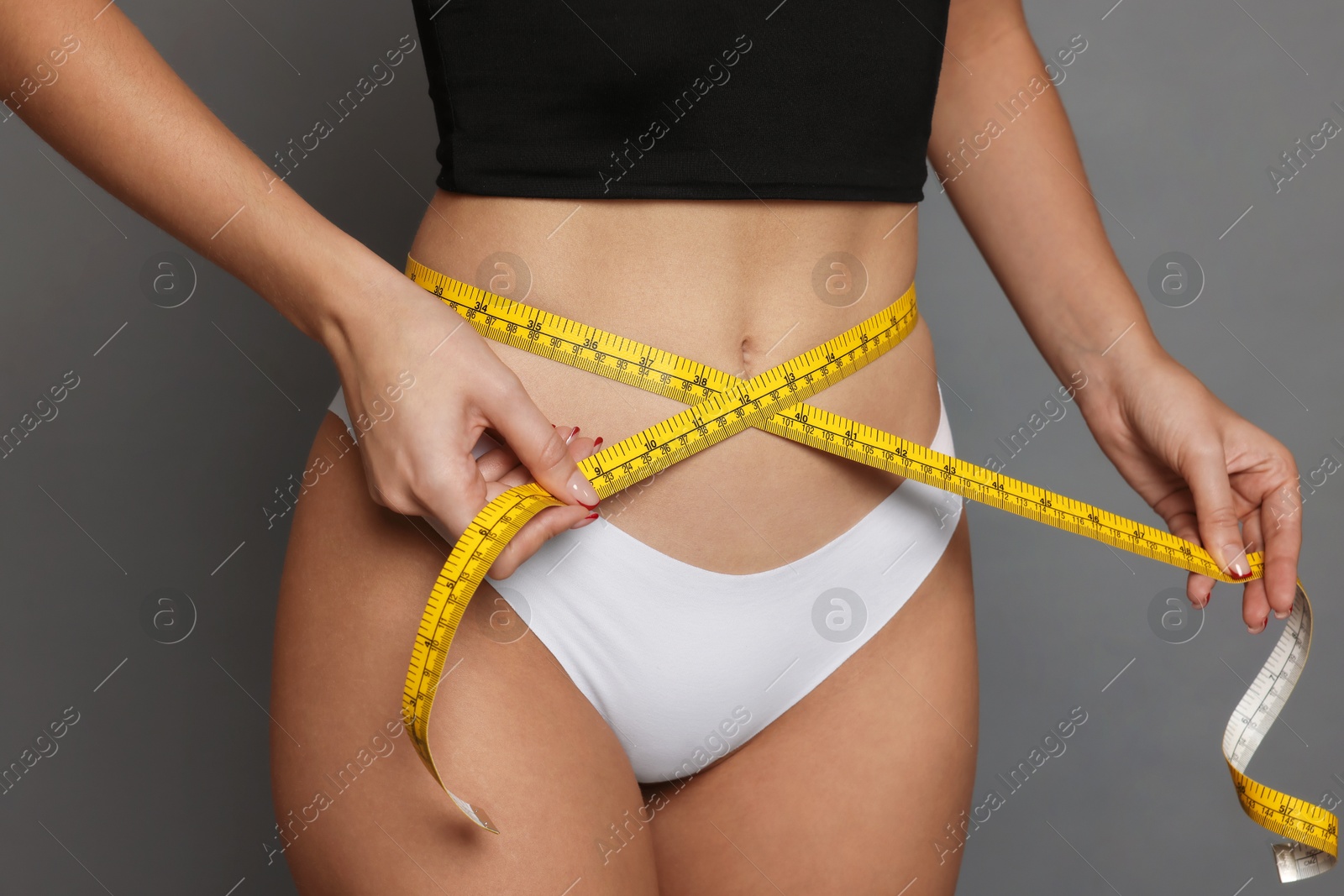 Photo of Young woman measuring her flat belly with tape on grey background, closeup