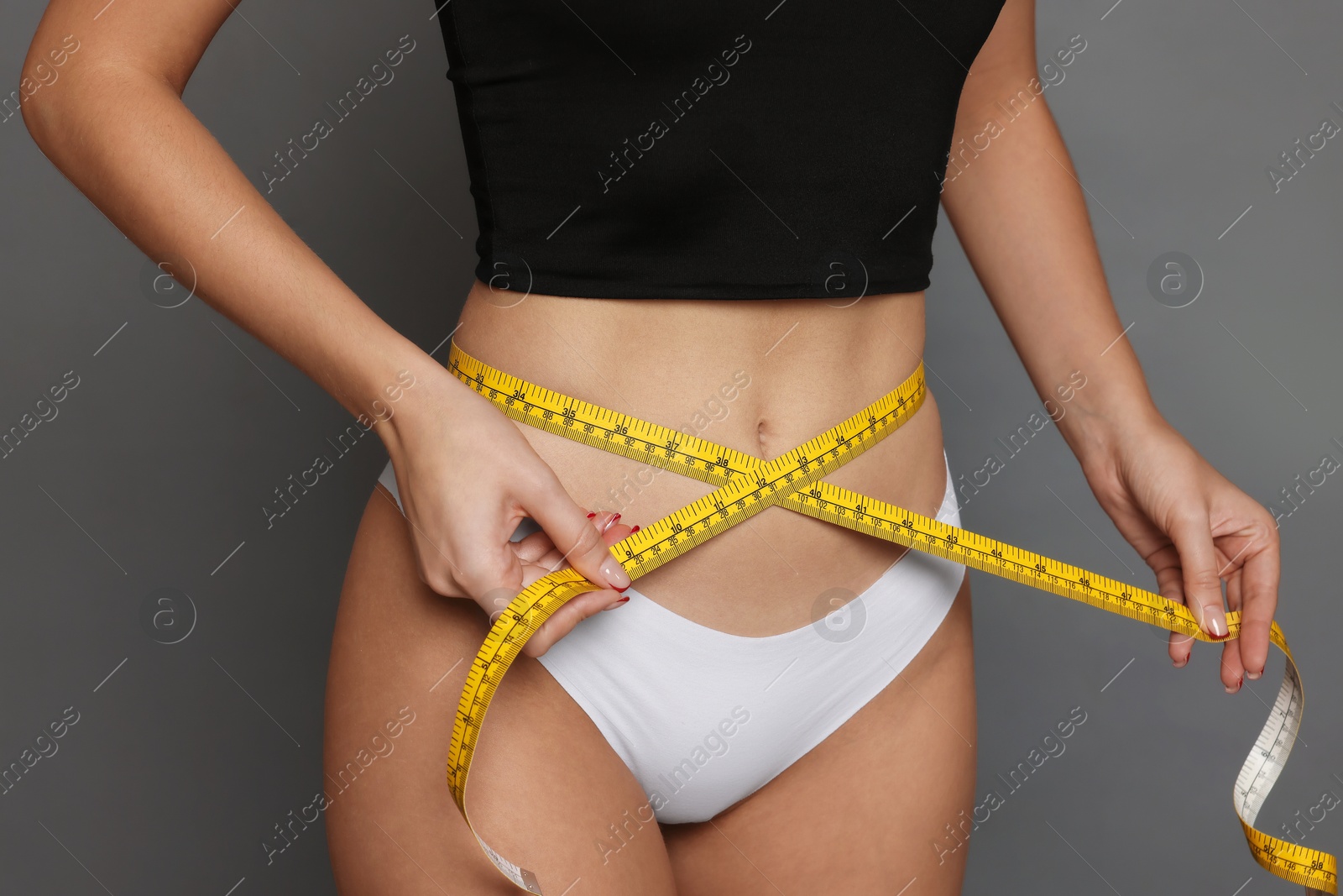 Photo of Young woman measuring her flat belly with tape on grey background, closeup
