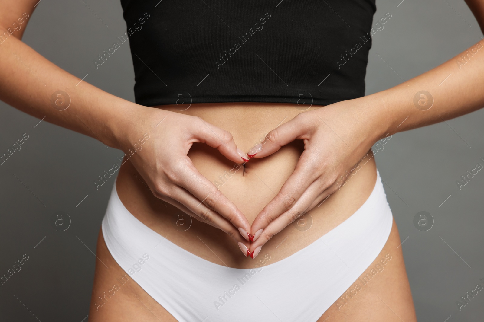 Photo of Young woman with flat belly making heart gesture on grey background, closeup