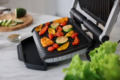 Photo of Electric grill with different vegetables on white table in kitchen, closeup