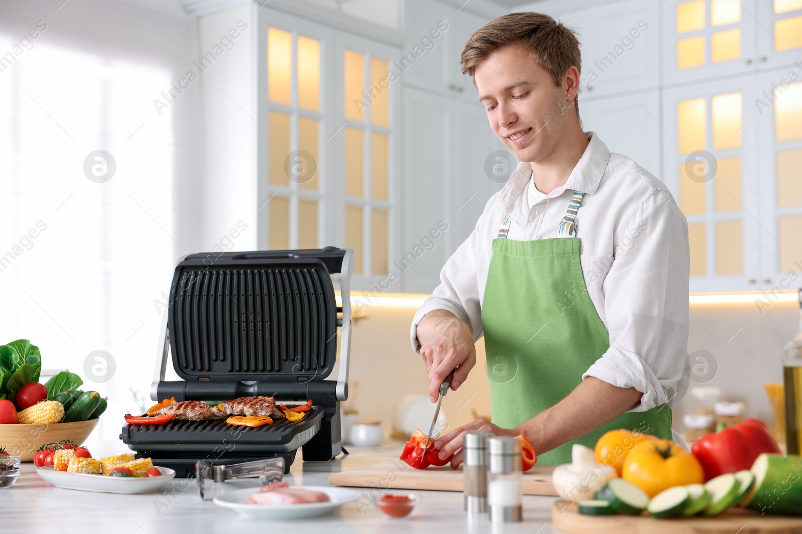 Photo of Man cutting pepper while cooking vegetables on electric grill in kitchen