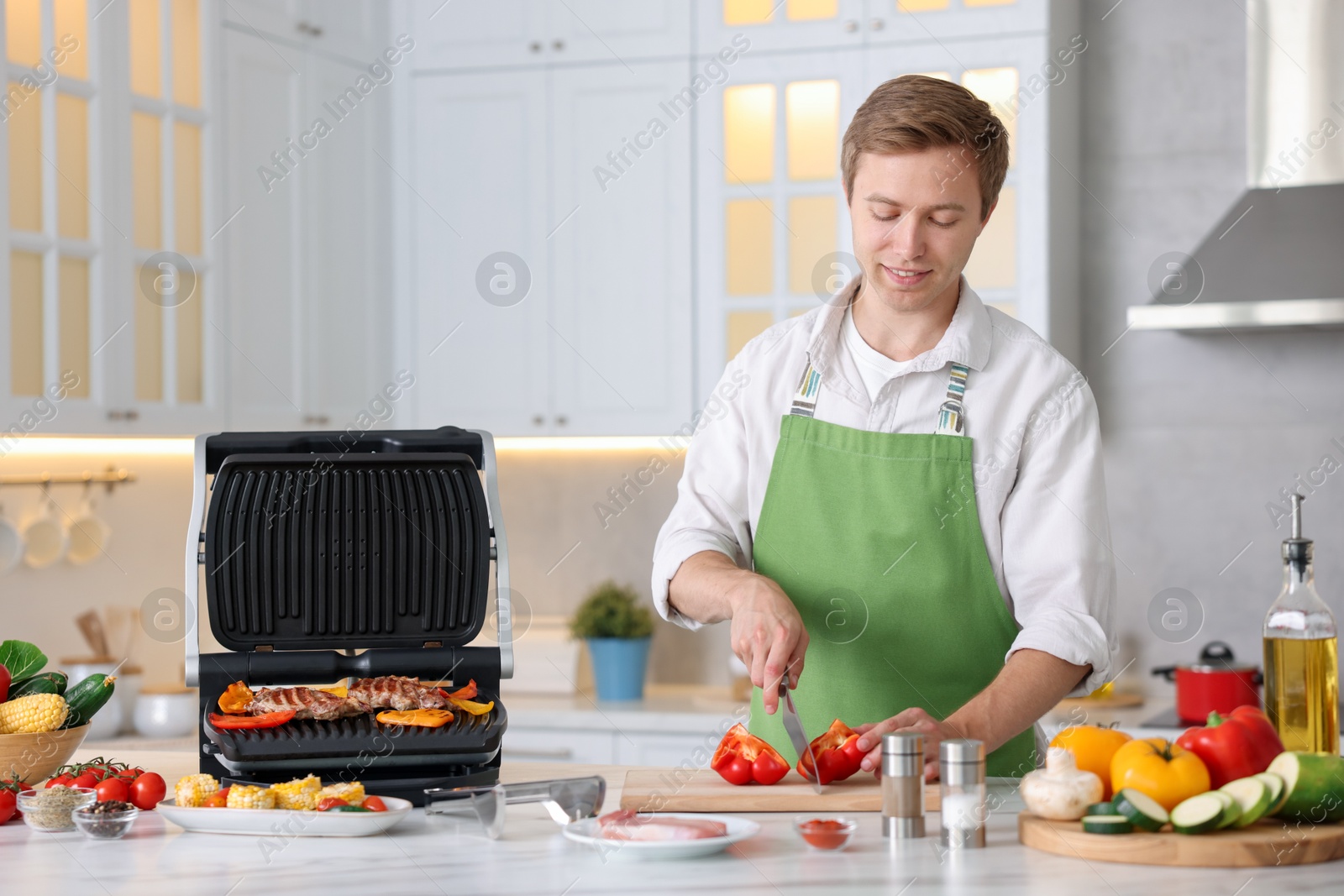 Photo of Man cutting pepper while cooking vegetables on electric grill in kitchen