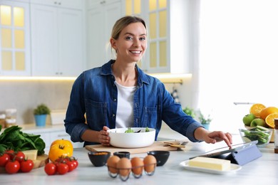 Photo of Smiling woman looking at recipe on tablet while cooking at table in kitchen