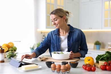 Photo of Smiling woman looking at recipe on tablet while cooking at table in kitchen