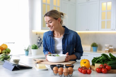 Photo of Smiling woman looking at recipe on tablet while cooking at table in kitchen