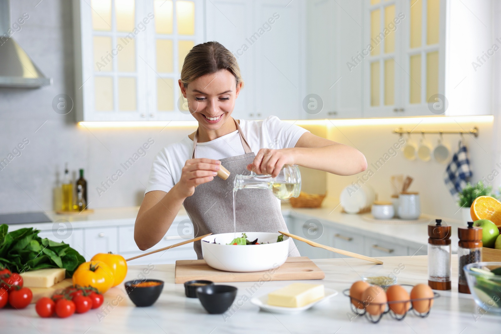 Photo of Cooking process. Smiling woman pouring oil onto salad at table in kitchen