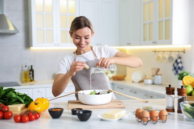 Photo of Cooking process. Smiling woman pouring oil onto salad at table in kitchen
