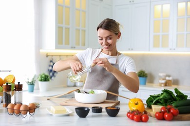 Photo of Cooking process. Smiling woman pouring oil onto salad at table in kitchen